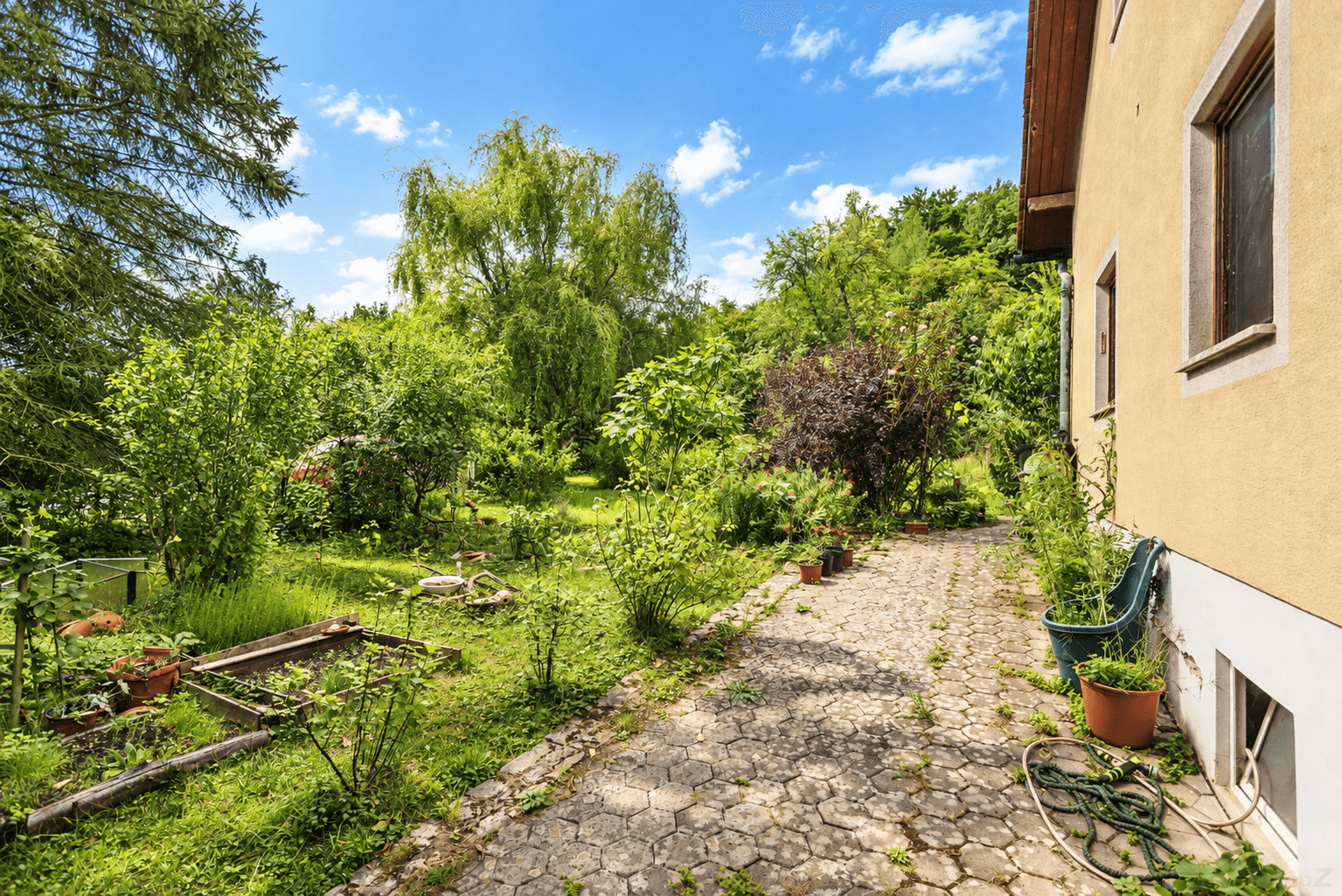 Gepflasterter Weg neben dem Haus, umgeben von einem üppigen, sonnigen Garten mit vielen Pflanzen.