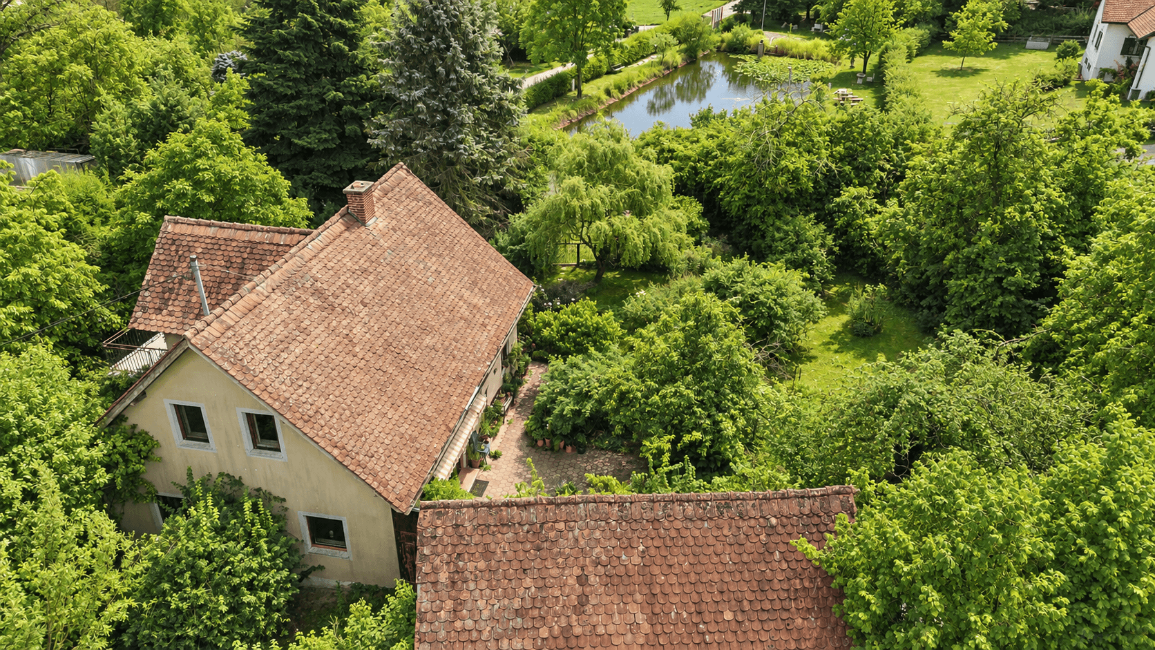 Luftaufnahme des Hauses und Gartens, umgeben von Bäumen, mit einem Teich in der Ferne sichtbar.