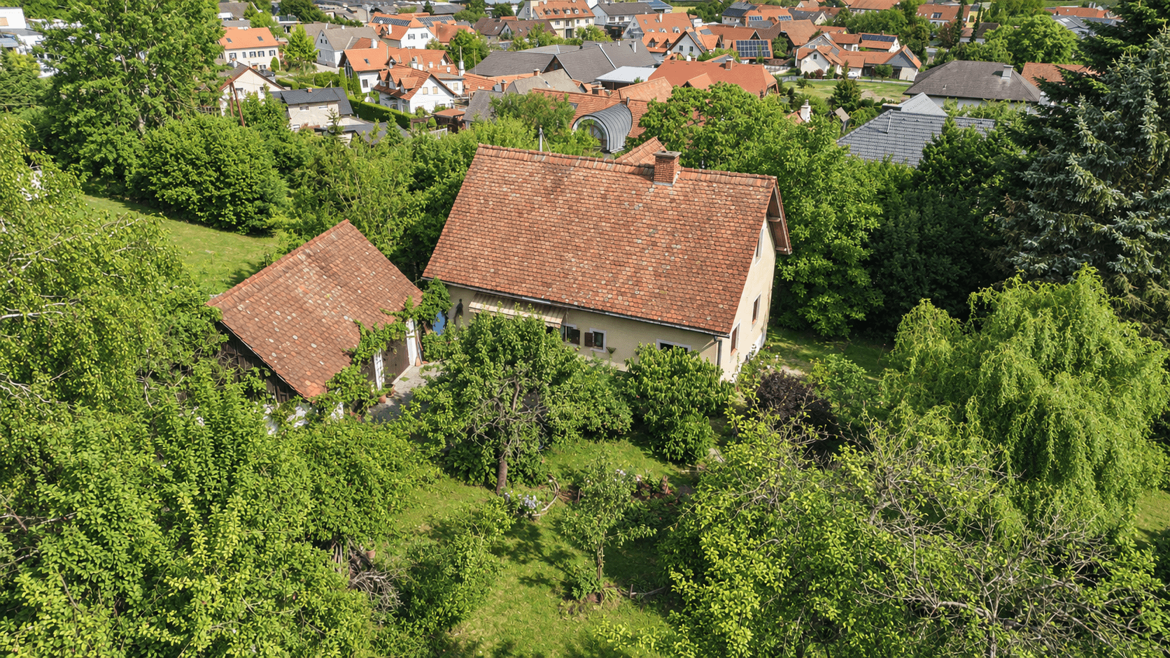 Luftaufnahme eines Hauses mit Nebengebäude, umgeben von viel Grün und einem Dorf im Hintergrund.