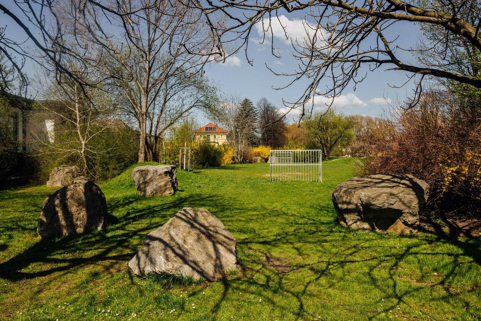 Großer Gartenbereich mit Rasenfläche, Felsen und einem kleinen Fußballtor unter Bäumen an einem sonnigen Tag.