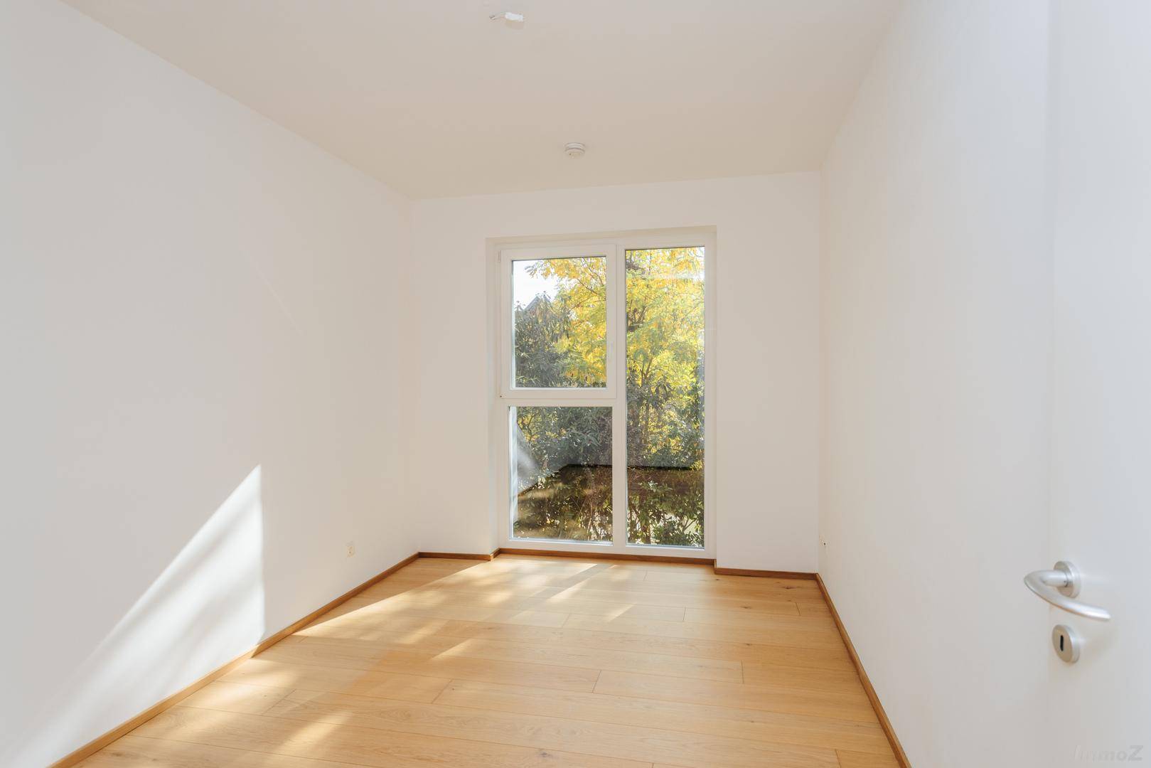 Leerer Raum mit hellem Holzboden und großem Fenster, das viel Tageslicht hereinlässt.