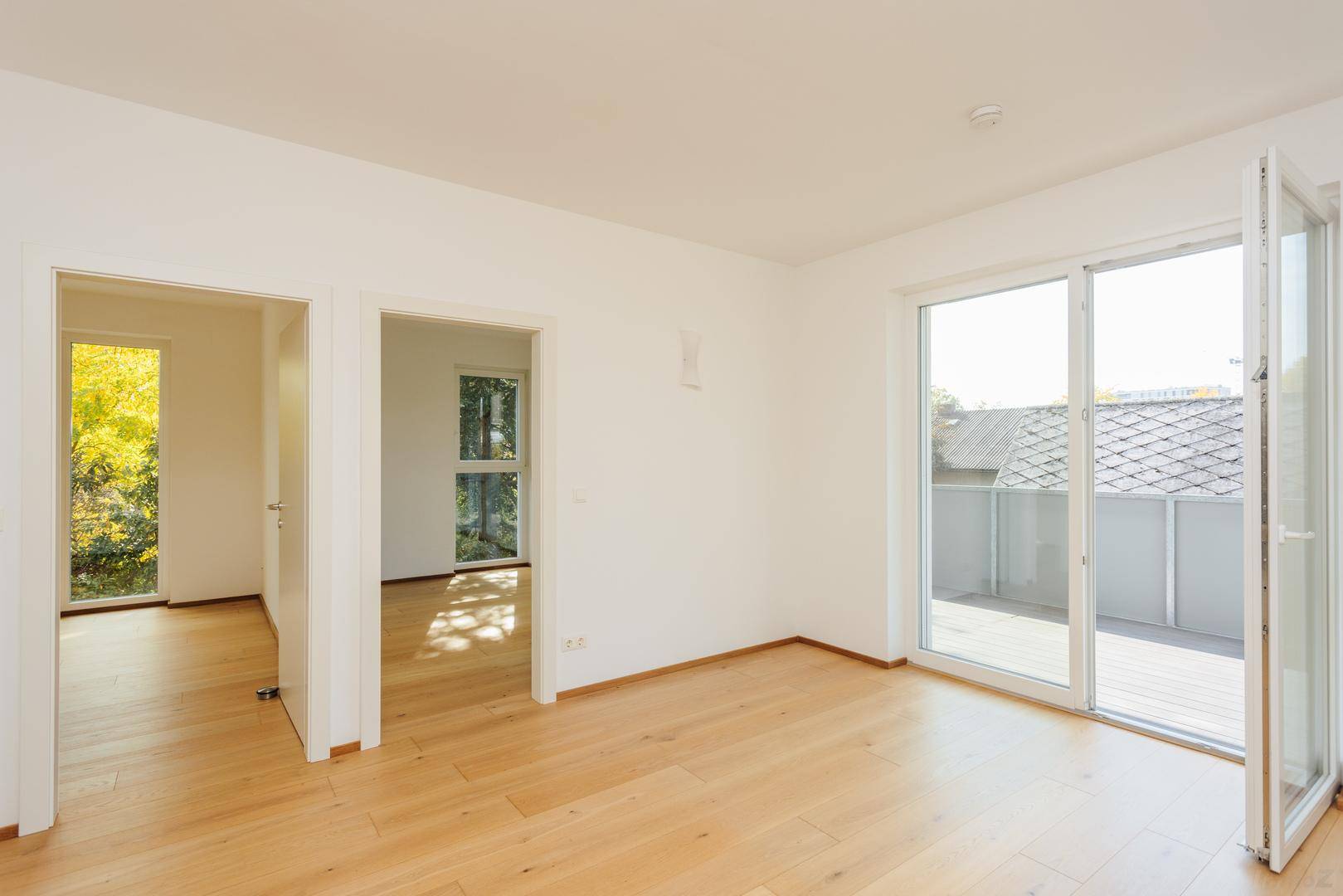 Geräumiger Raum mit Holzboden, mehreren Türen und direktem Zugang zu einem sonnigen Balkon.