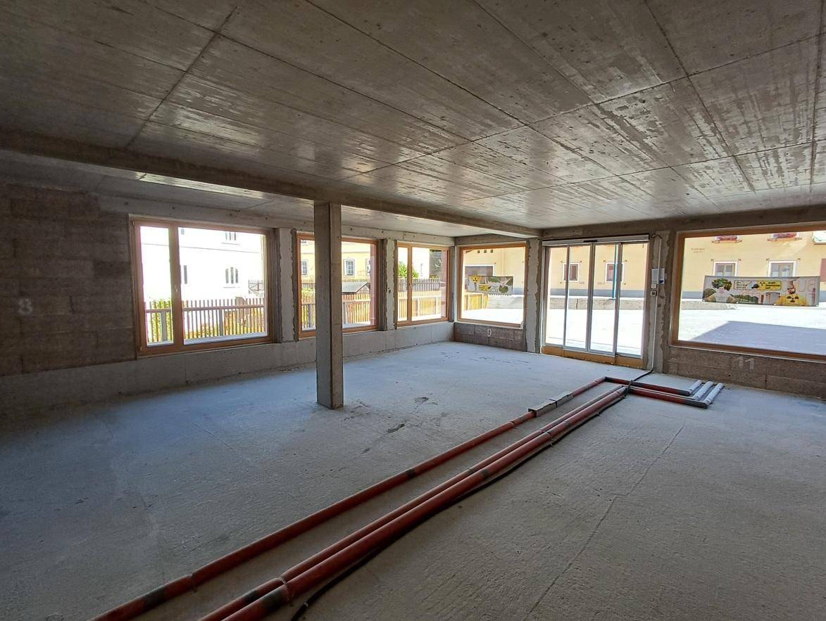 Großer Innenraum im Rohbau mit vielen Fenstern, einer Schiebetür und sichtbaren Installationen am Boden.