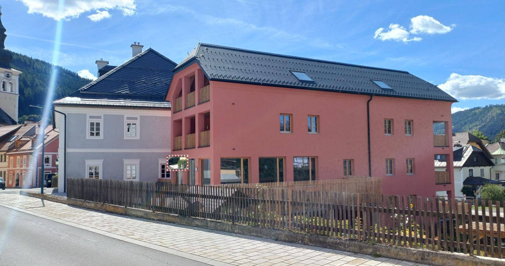 Seitenansicht des modernen Gebäudes mit rosafarbener Fassade, dunklem Dach und Holzzaun.