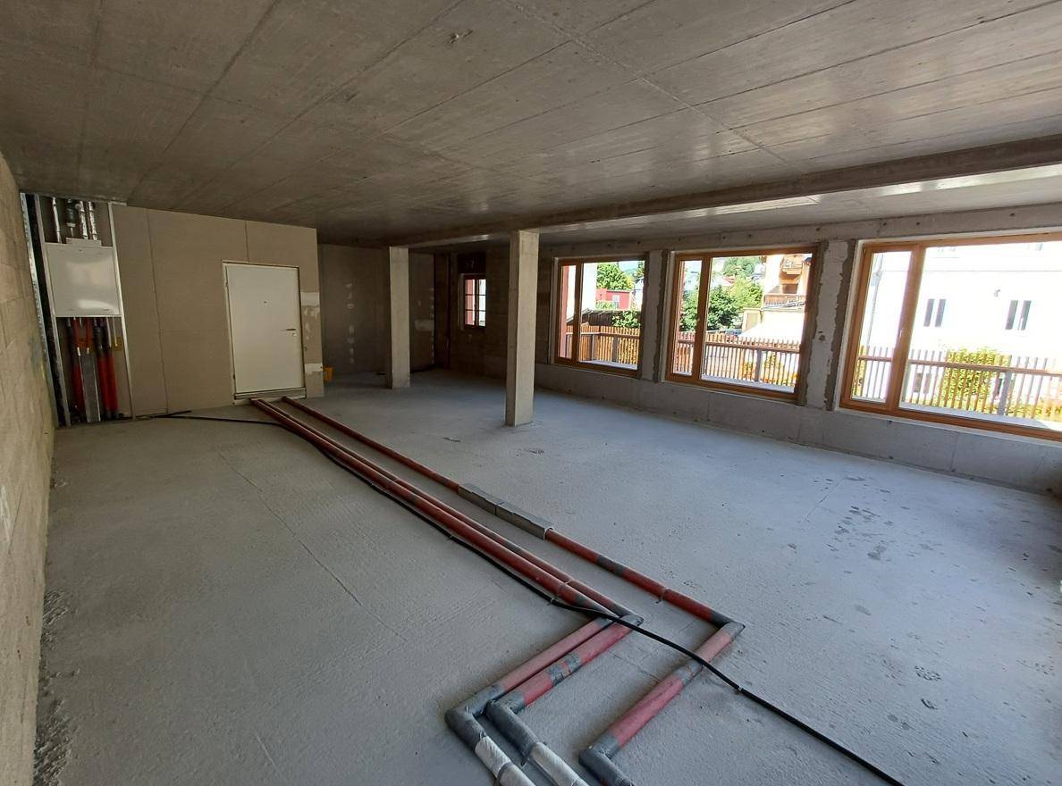 Unfertiger Innenraum mit Betonboden, großen Fenstern, einer Tür und sichtbaren Rohrleitungen an der Wand.
