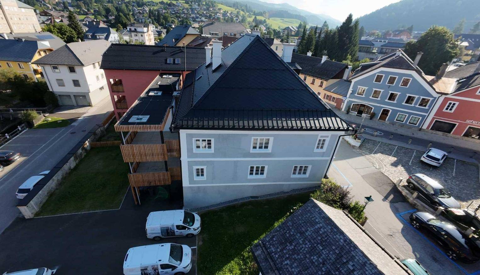 Luftaufnahme des Anwesens mit umliegenden Häusern, Parkplätzen und einer bergigen Landschaft im Hintergrund.