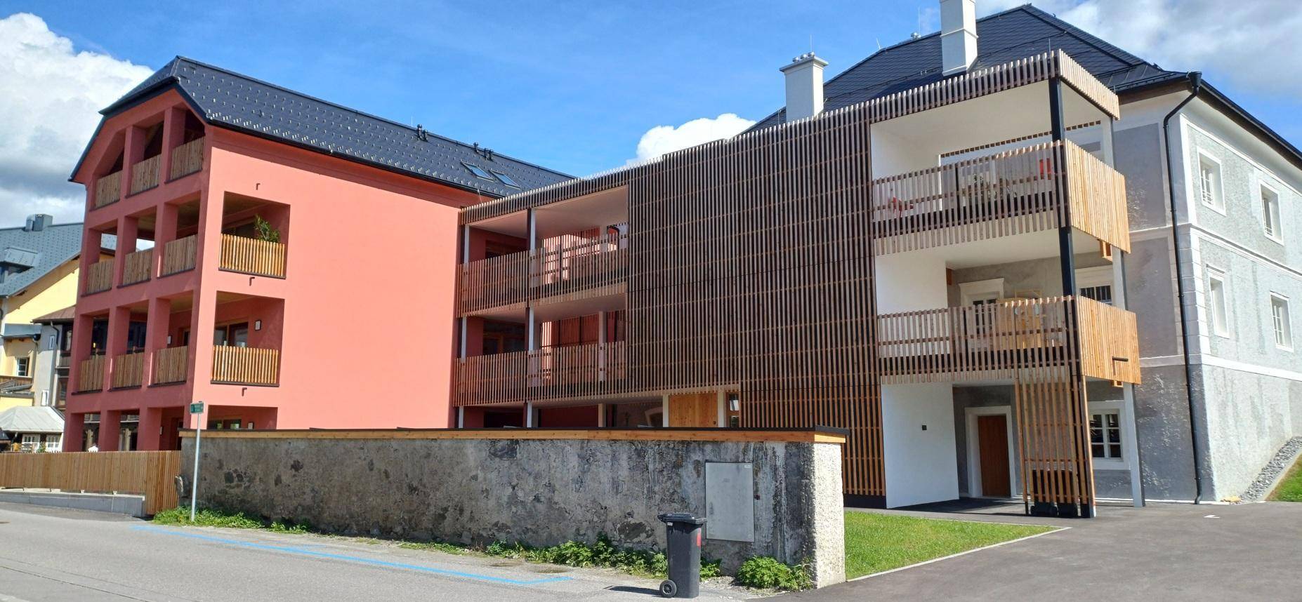 Moderne und traditionelle Gebäude nebeneinander, mit Holzelementen und Balkonen am Neubau, unter blauem Himmel.
