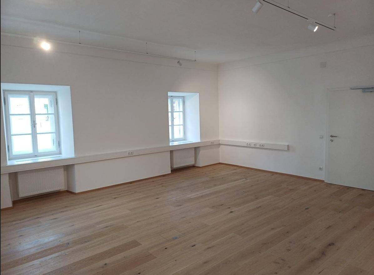 Heller, leerer Raum mit Holzboden, weißen Wänden und zwei Fenstern mit Heizkörpern darunter, ideal für Büro oder Wohnen.