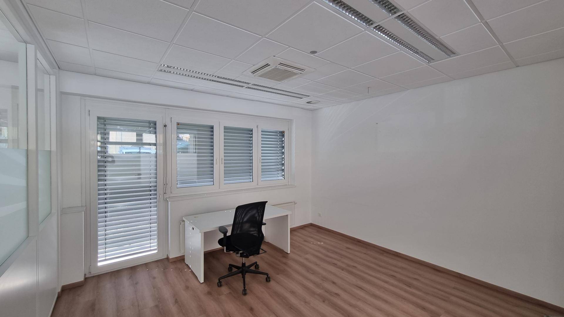 Helles Einzelbüro mit Schreibtisch, Bürostuhl, Jalousien an den Fenstern und Laminatboden.