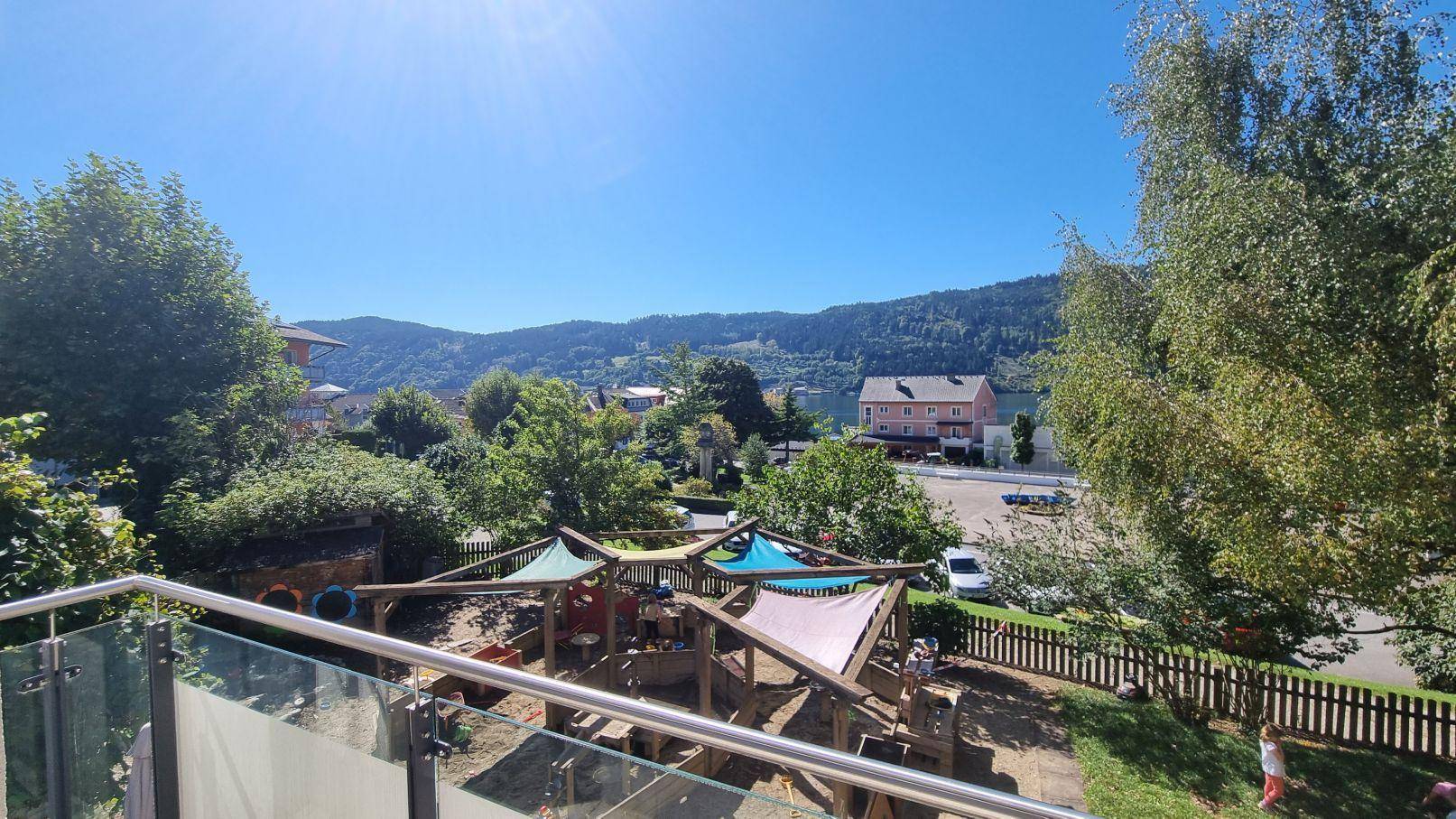 Sonniger Balkon mit Blick auf einen Spielplatz, grüne Bäume und eine malerische Berglandschaft im Hintergrund.