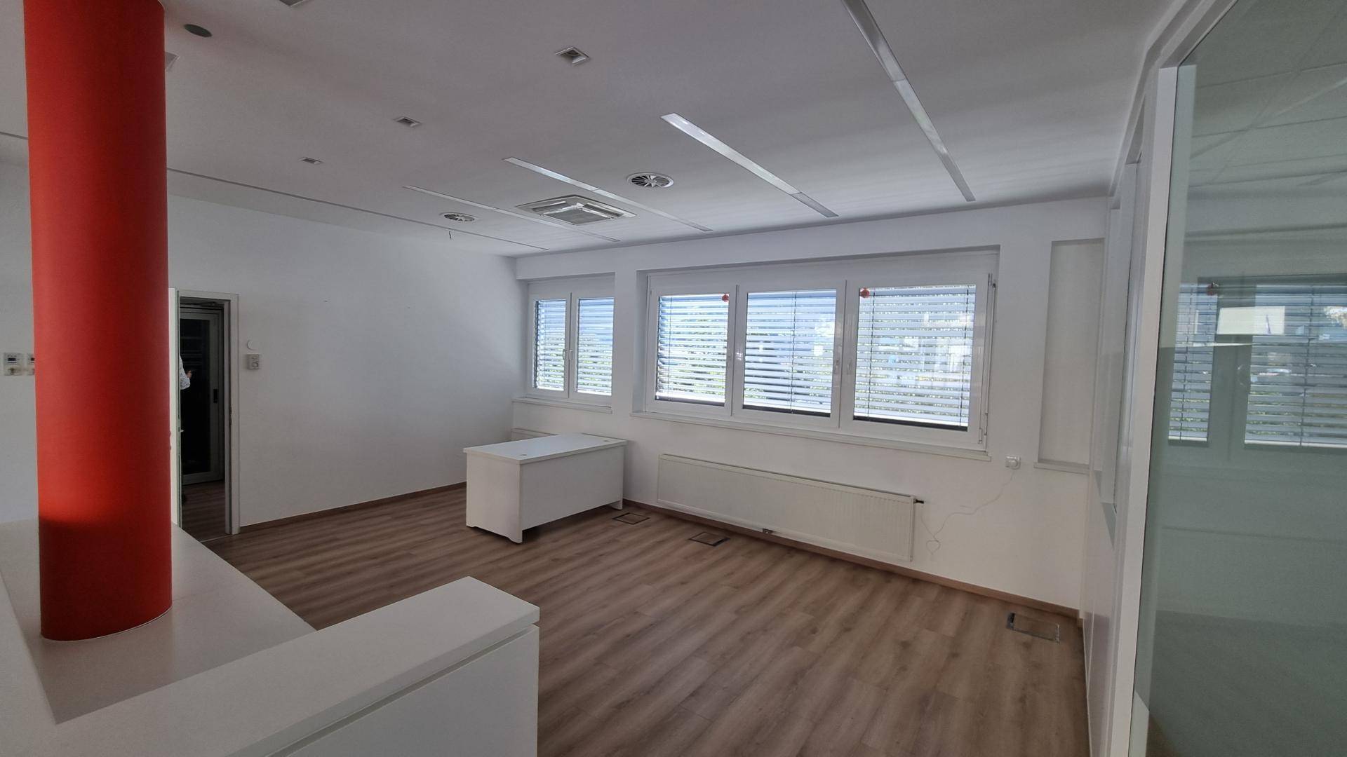 Geräumiger Bürobereich mit Laminatboden, Heizkörpern unter den Fenstern und einer auffälligen roten Säule.