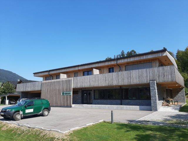 Modernes Gebäude mit Holzfassade und Steinelementen, umgeben von Grünflächen und Parkplätzen.