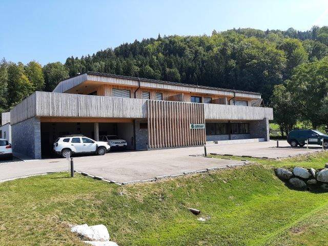 Ansicht des modernen Gebäudes mit Holzfassade, Steinsockel und großzügigen Parkmöglichkeiten im Grünen.