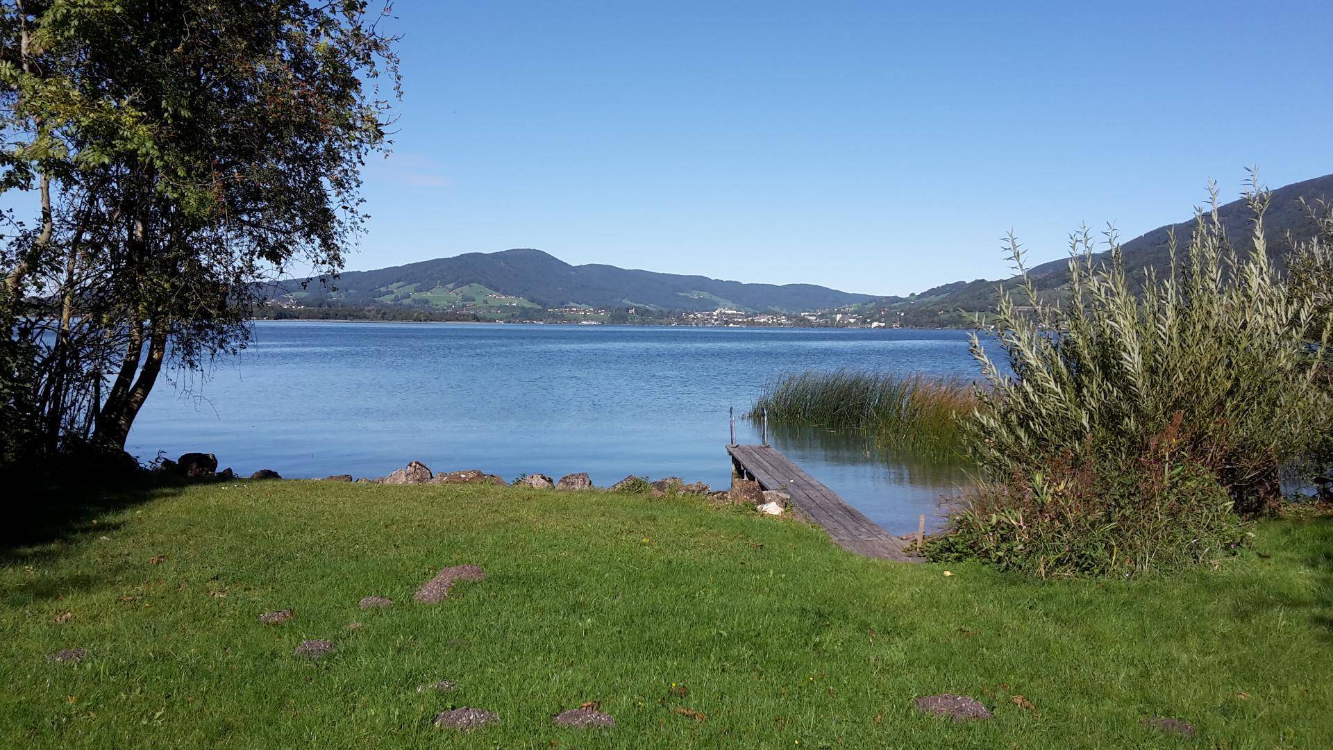 Ruhiger See mit kleinem Holzsteg, umgeben von grünen Hügeln und Bäumen unter blauem Himmel.