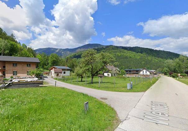 Ländliche Straße führt durch eine Siedlung mit Häusern und grünen Wiesen vor einer Bergkulisse.