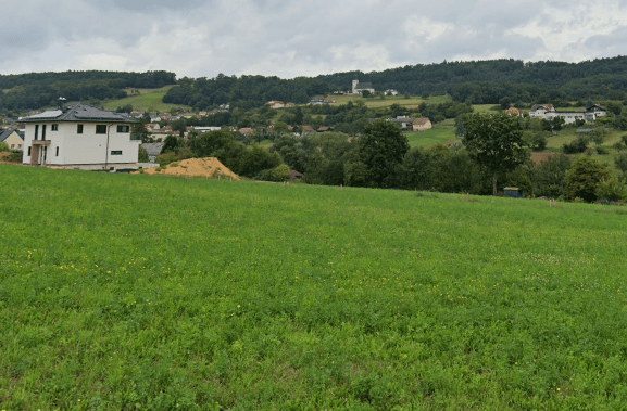 Weitläufige grüne Wiesen mit einem modernen Haus im Vordergrund und einem Dorf am Hügel im Hintergrund.