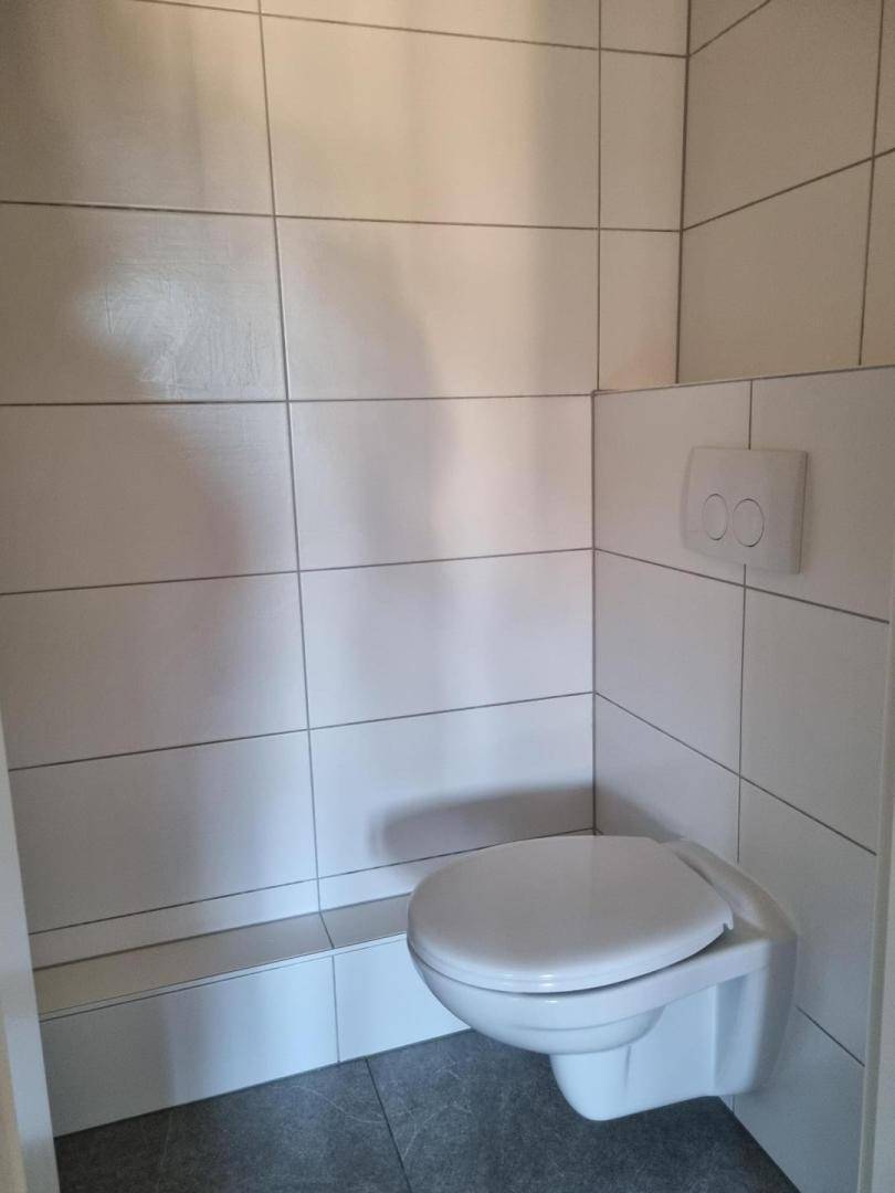 Separates WC mit wandhängender Toilette und weißen Wand- und Bodenfliesen.