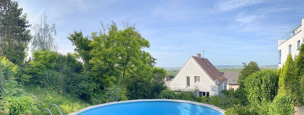 Panoramablick auf den Außenpool mit weitreichender grüner Landschaft und Häusern im Hintergrund.