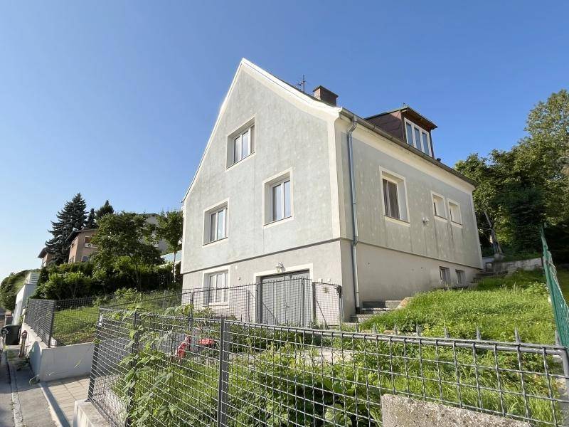 Seitenansicht des Hauses mit heller Fassade, Ziegeldach und Hanglage im Grünen.