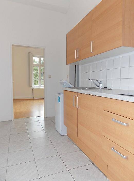 Einfache Küche mit Holzschränken, Fliesenboden und Durchgang zu einem weiteren Raum.