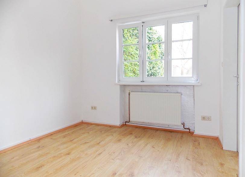 Leeres Zimmer mit Laminatboden, weißen Wänden und Fenster mit Blick ins Grüne.