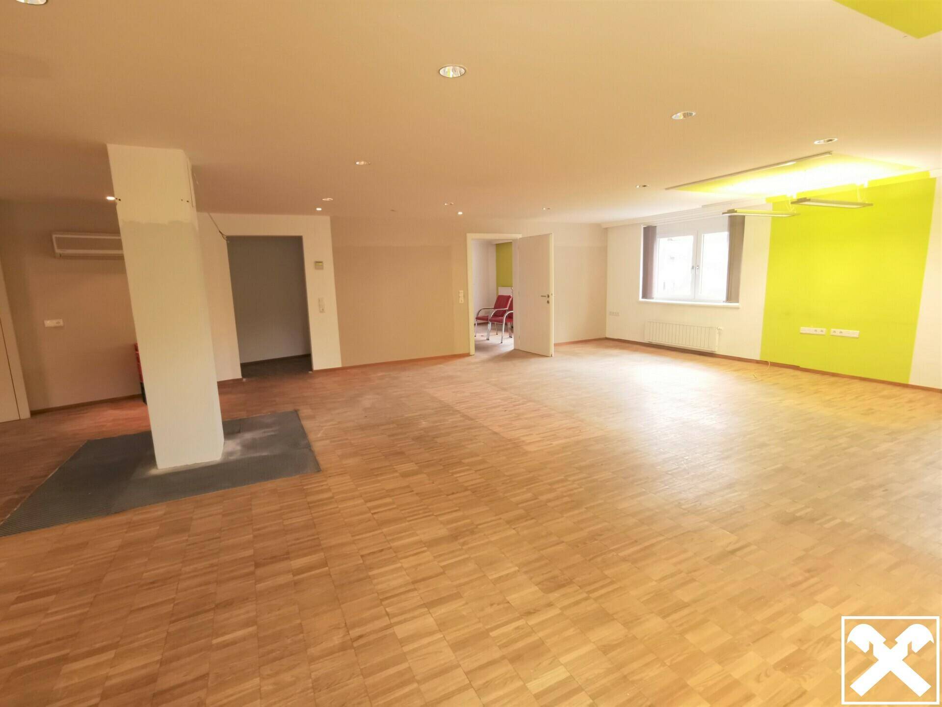 Weitläufiger Bürobereich mit Holzboden, Säule in der Mitte und Zugang zu weiteren Räumen.