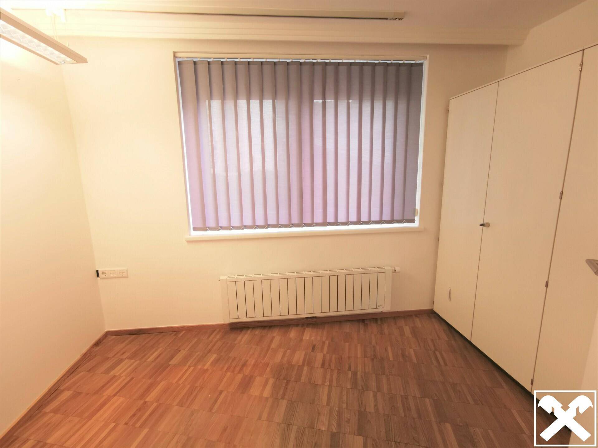 Kleinerer Büroraum mit Holzparkett, Einbauschrank und Fenster mit vertikalen Jalousien.