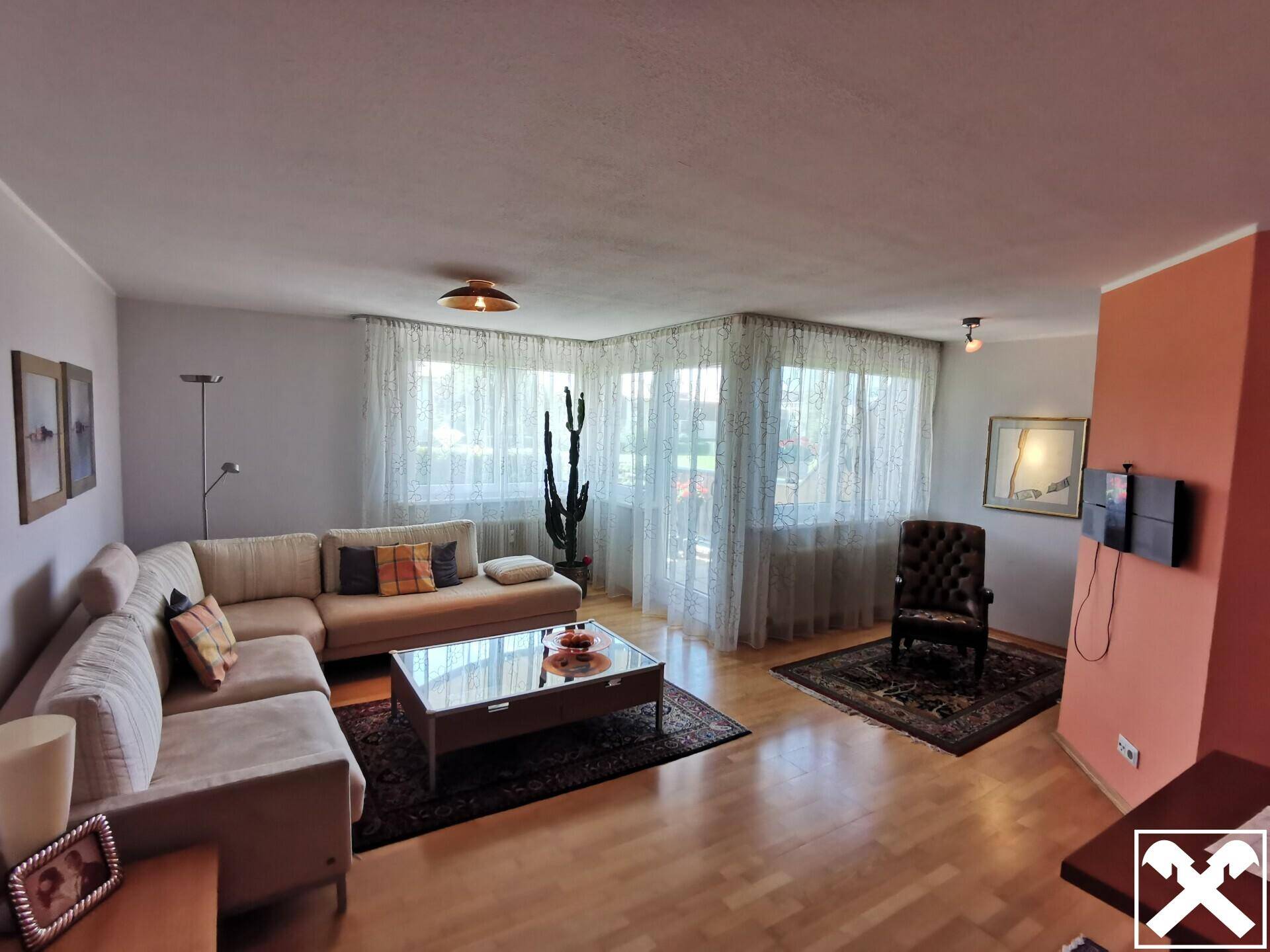 Gemütliches Wohnzimmer mit Ecksofa, großen Fenstern und hellem Holzboden, das viel Tageslicht hereinlässt.
