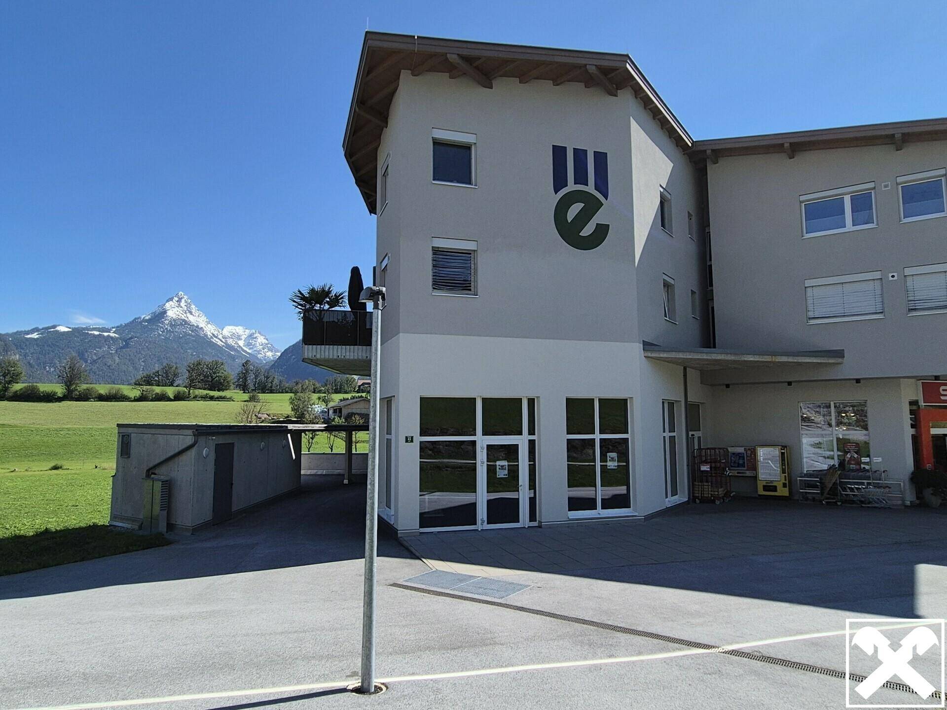 Modernes Gebäude mit großen Fenstern und Balkon, umgeben von grünen Feldern und beeindruckendem Bergblick.