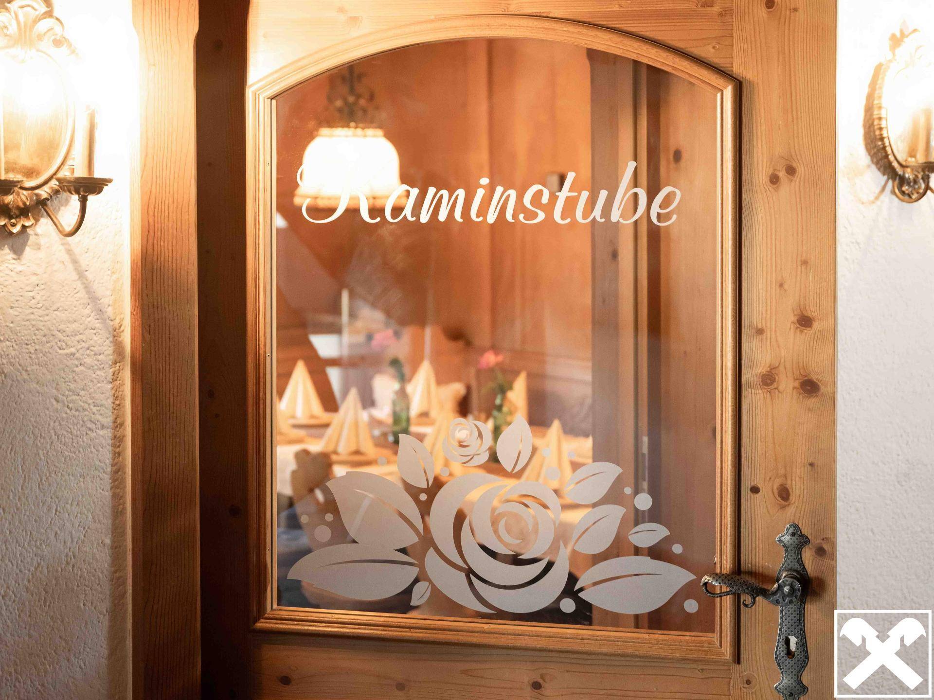 Holztür mit Glaseinsatz und "Kaminstube"-Schriftzug, die einen Blick in einen traditionell gedeckten Essbereich freigibt.
