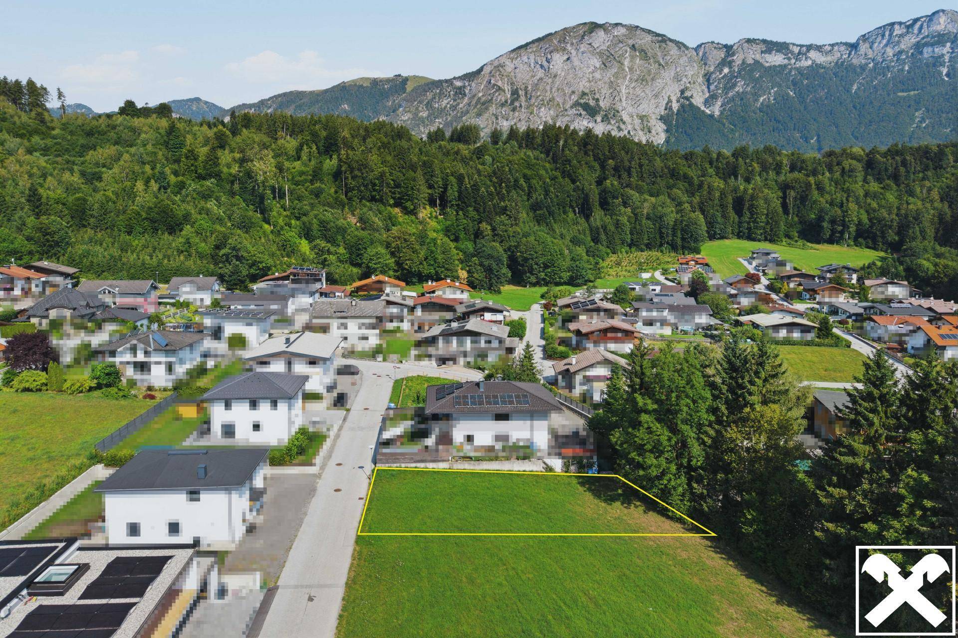 Luftaufnahme einer Wohnsiedlung mit markiertem Grundstück, umgeben von Natur und Bergen.