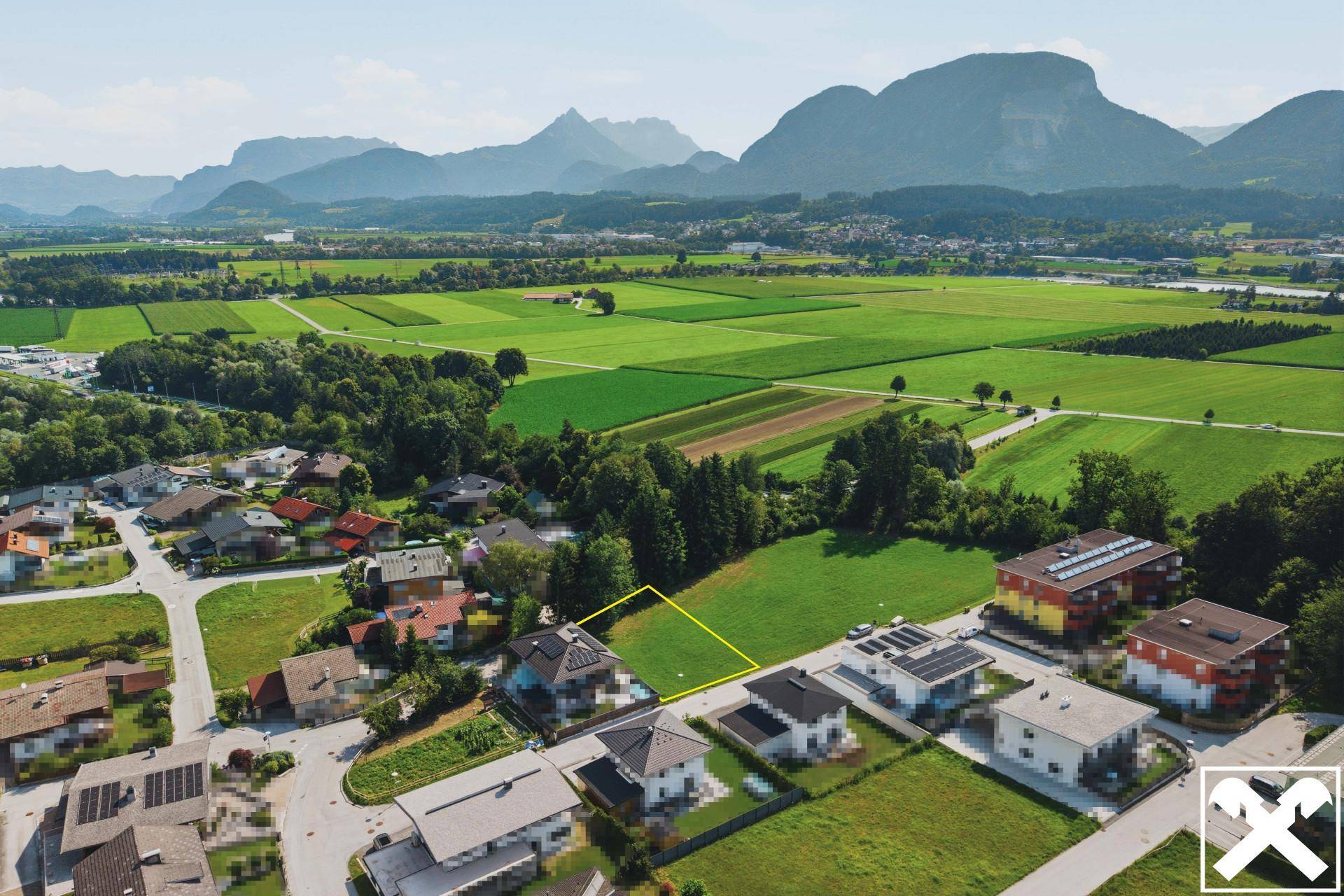 Luftaufnahme einer Wohnsiedlung mit weiten Feldern, Bergen im Hintergrund und einem markierten Grundstück.