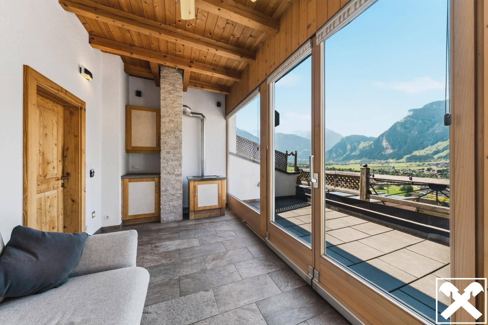 Heller Wohnbereich mit großen Fenstern, Holzbalkendecke und Zugang zu einer sonnigen Terrasse mit Bergblick.