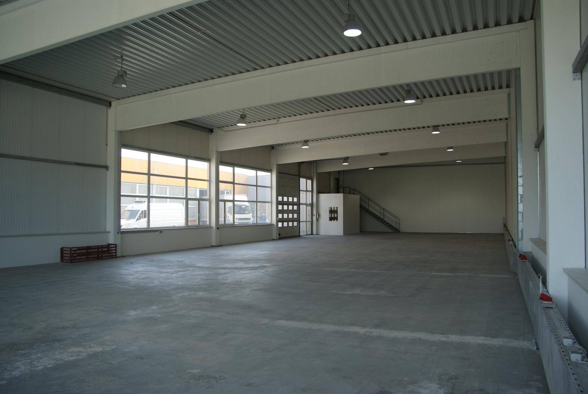 Geräumige, leere Lagerhalle mit vielen großen Fenstern, die viel Licht hereinlassen, und einem Rolltor.