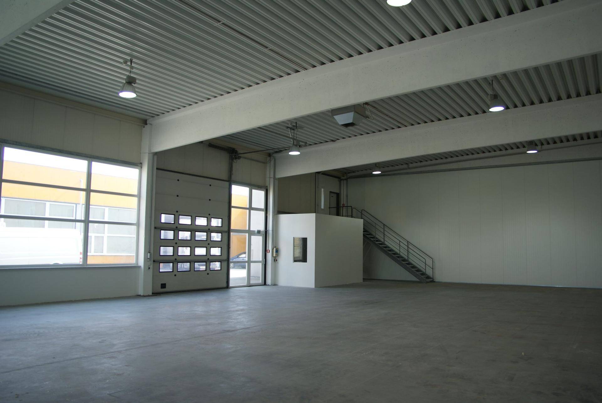 Innenansicht der Lagerhalle mit Rolltor, großen Fenstern, einer Metalltreppe und einem kleinen, abgetrennten Raum.