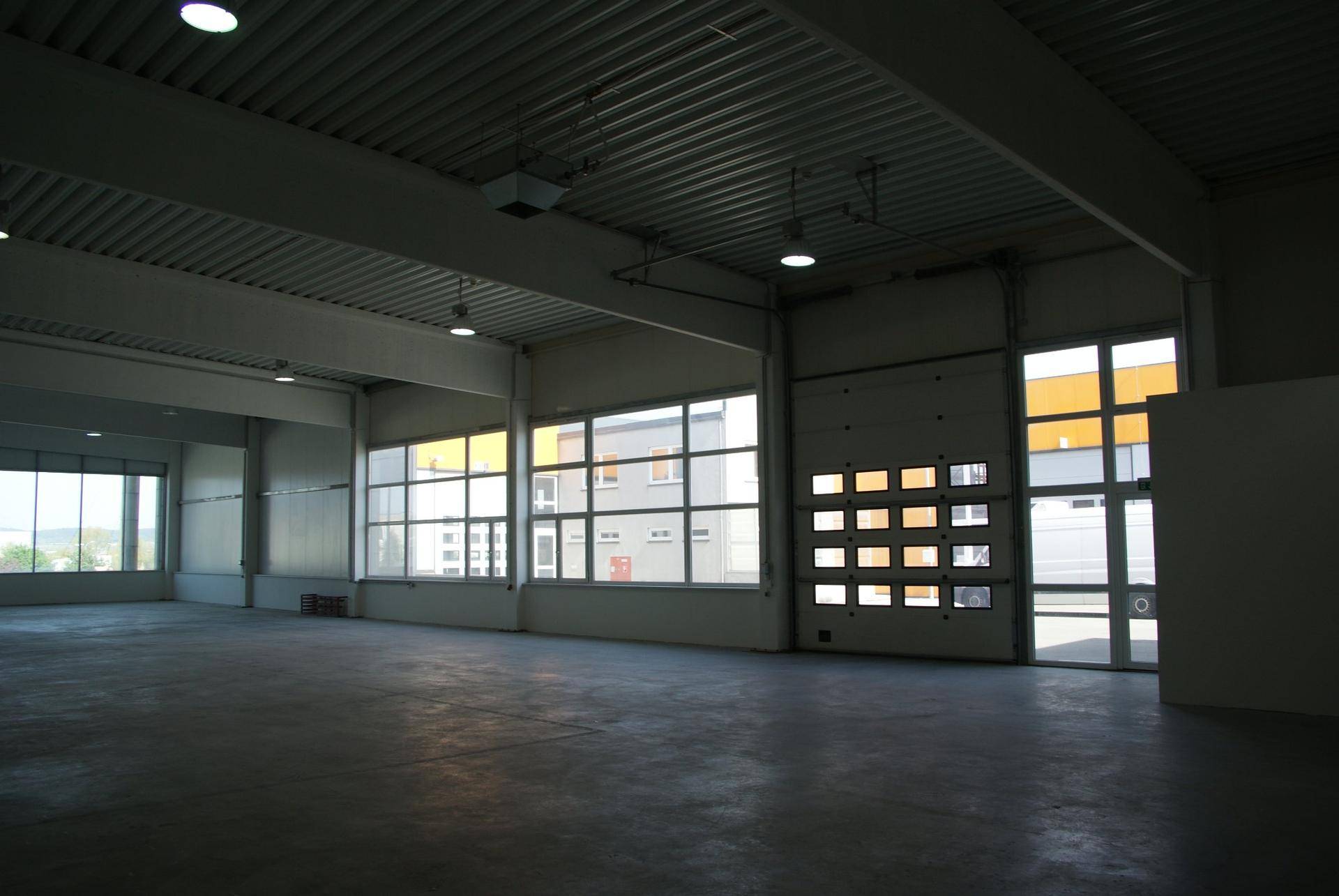 Weitläufige Lagerhalle mit Betonboden, hohen Decken und einem großen Rolltor mit integrierten Fenstern.