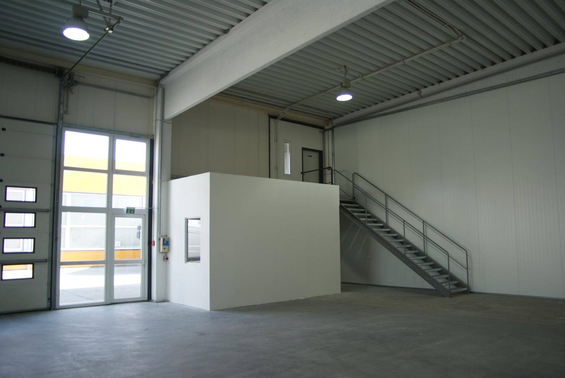 Große Lagerhalle mit hohen Decken, Betonboden, einer Metalltreppe und einem kleinen, abgetrennten Bereich.