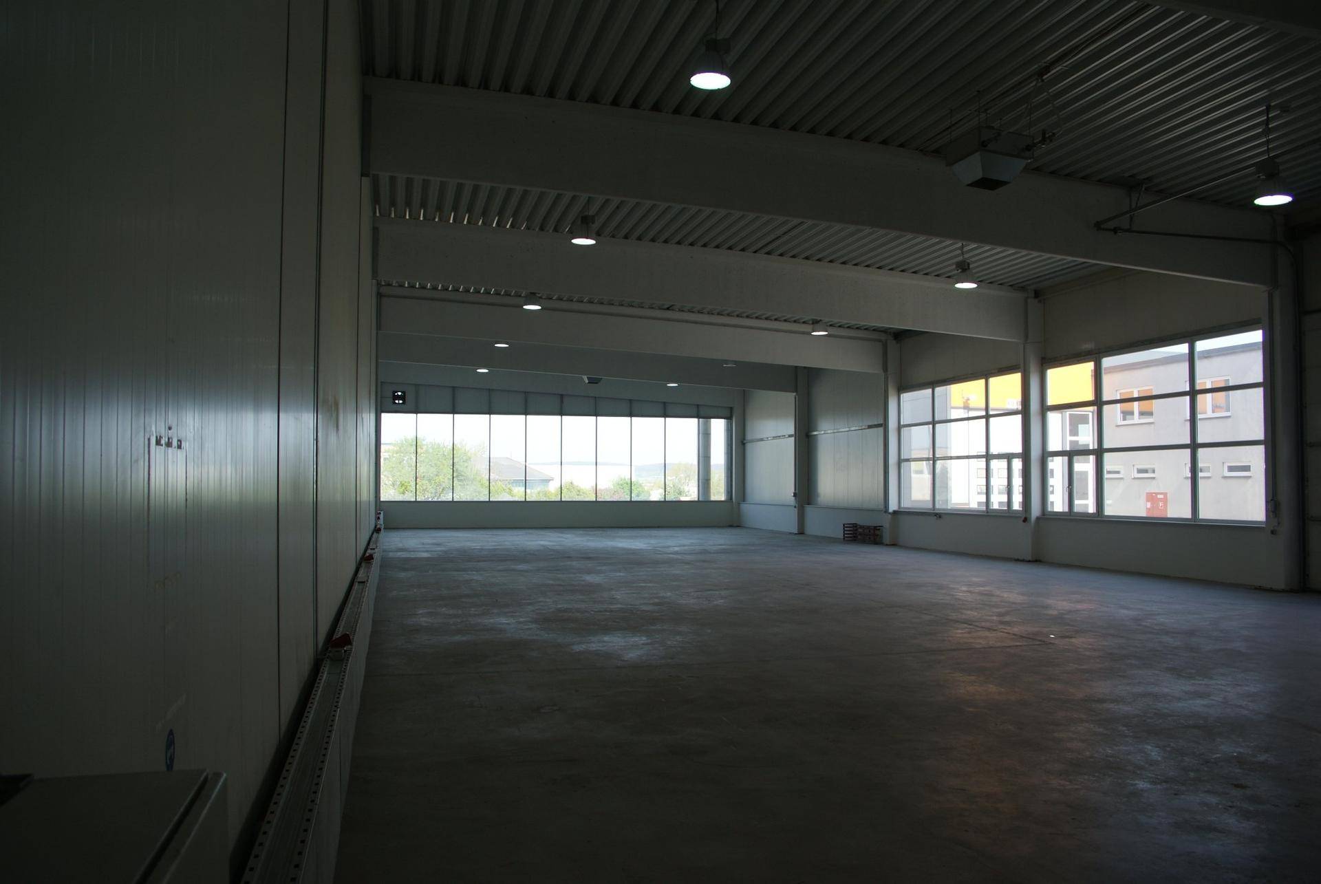 Geräumige, leere Lagerhalle mit hohen Decken, Betonboden und großen Fenstern, die viel Tageslicht hereinlassen.