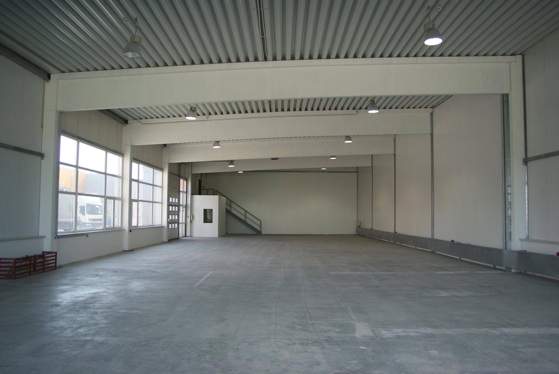 Weitläufige, leere Lagerhalle mit hohen Decken, Betonboden und großen Fensterfronten für natürliche Belichtung.