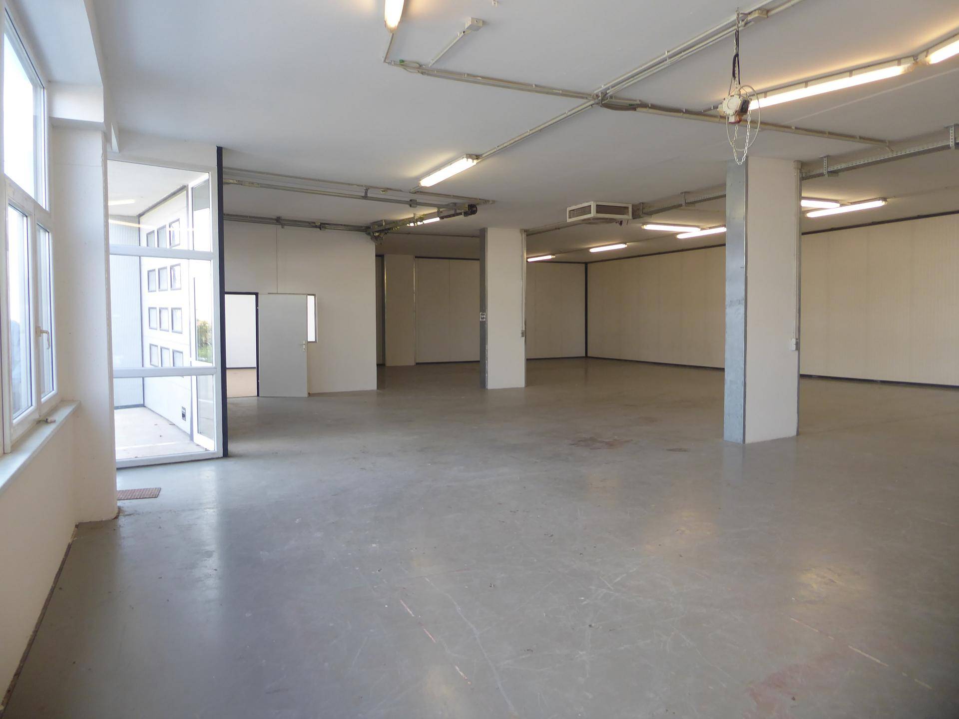 Geräumiger, heller Lagerraum mit großen Fenstern, Betonboden und sichtbaren Installationen an der Decke.