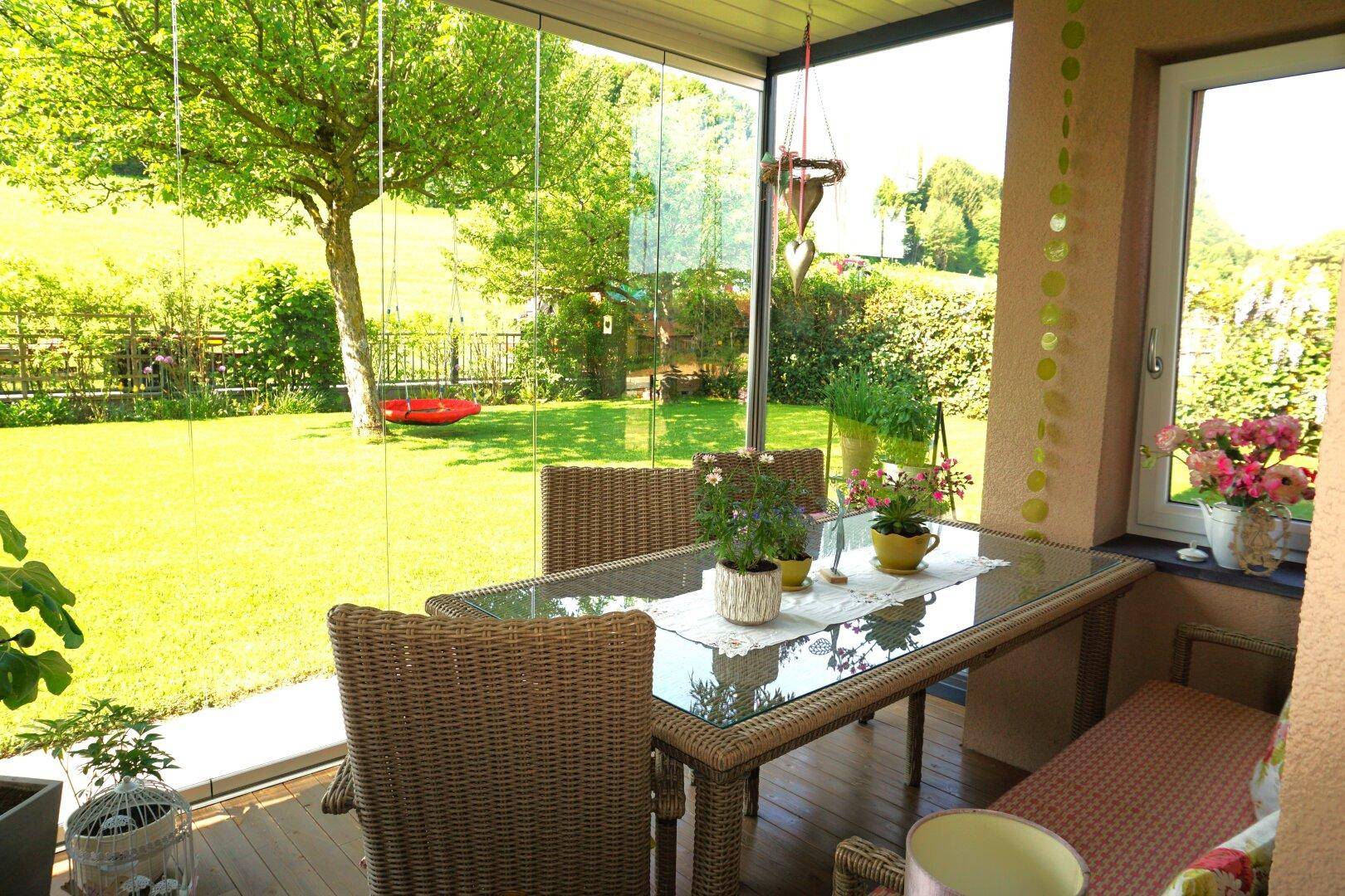 Heller Wintergarten mit Esstisch, Korbstühlen und weitem Blick ins Grüne.