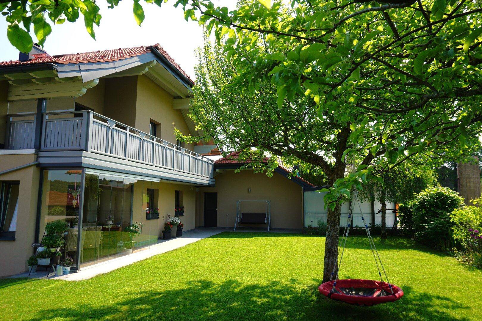 Hausfassade mit großem grünen Garten, Schaukel unter einem Baum und verglastem Wintergarten.