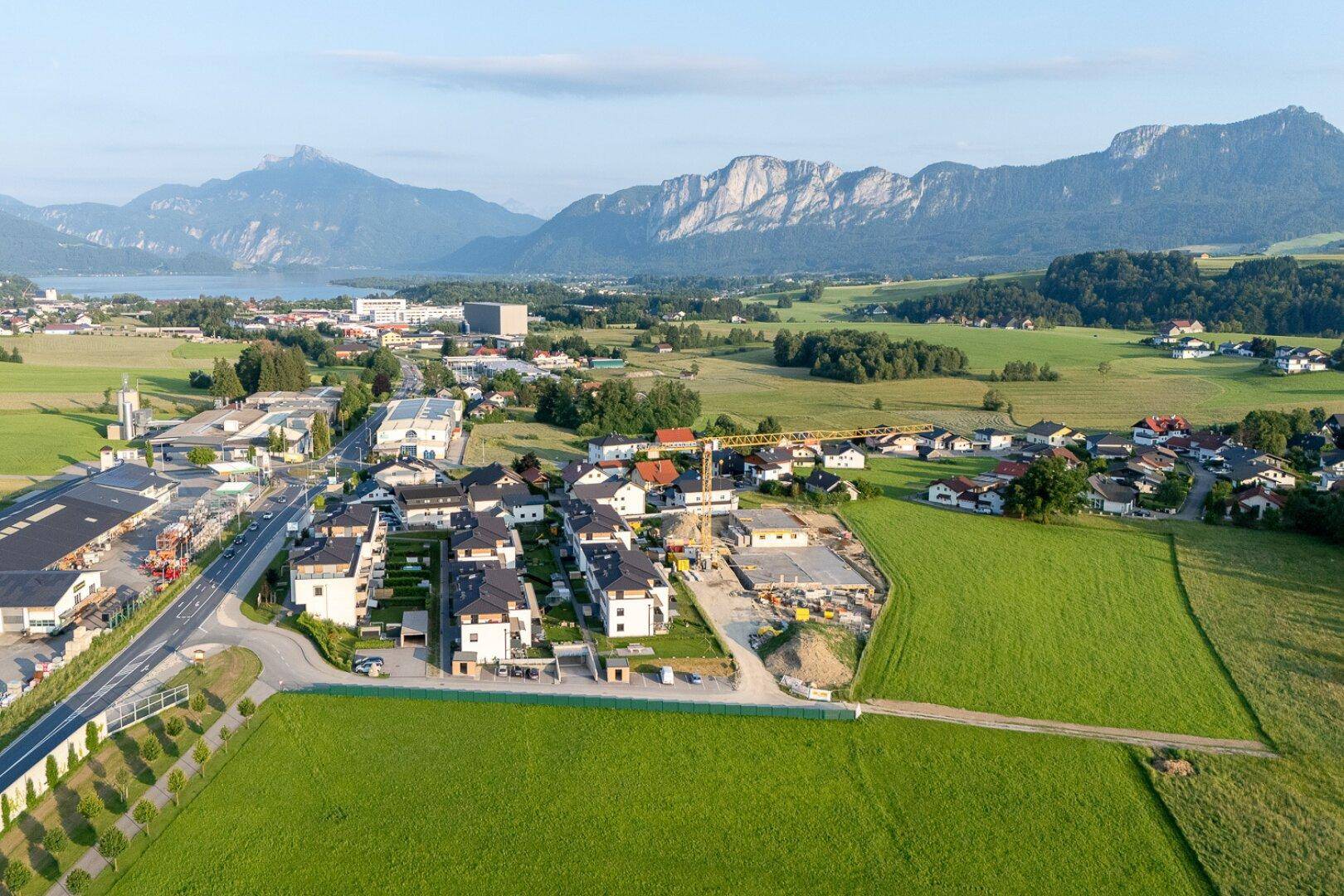Luftaufnahme einer Wohnsiedlung mit Neubauten und Baustelle, umgeben von grüner Landschaft und Bergen.