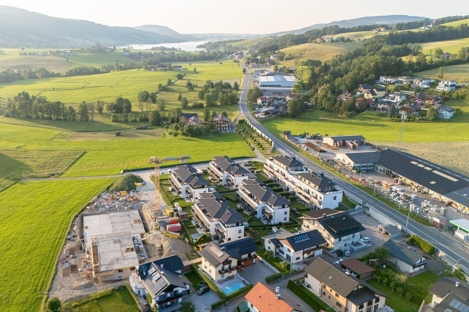 Luftaufnahme einer Wohnsiedlung mit Neubauten und Baustelle, umgeben von grüner Landschaft und Bergen.