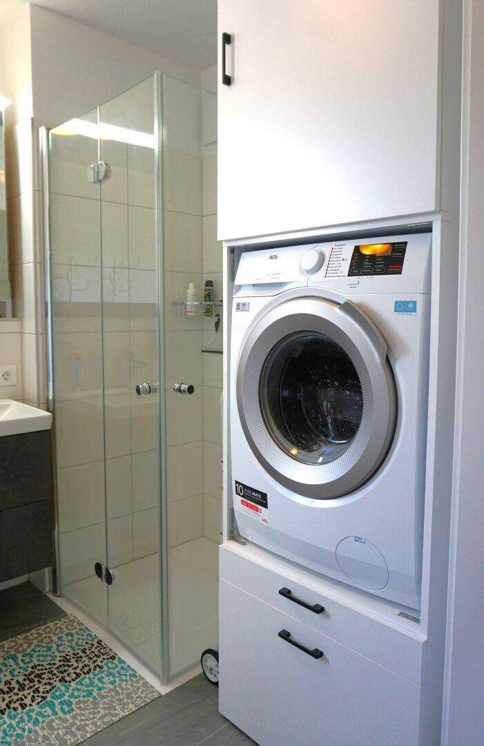 Integrierte Waschmaschine und Trockner in einem weißen Schrank im Badezimmer, neben einer Duschkabine.