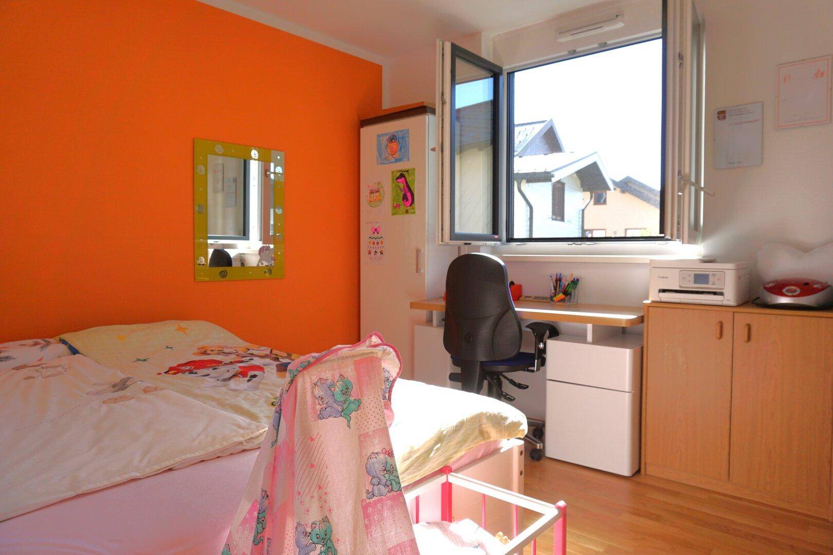 Kinderzimmer mit leuchtend orangefarbener Wand, Holzboden und einem Schreibtisch am großen Fenster.