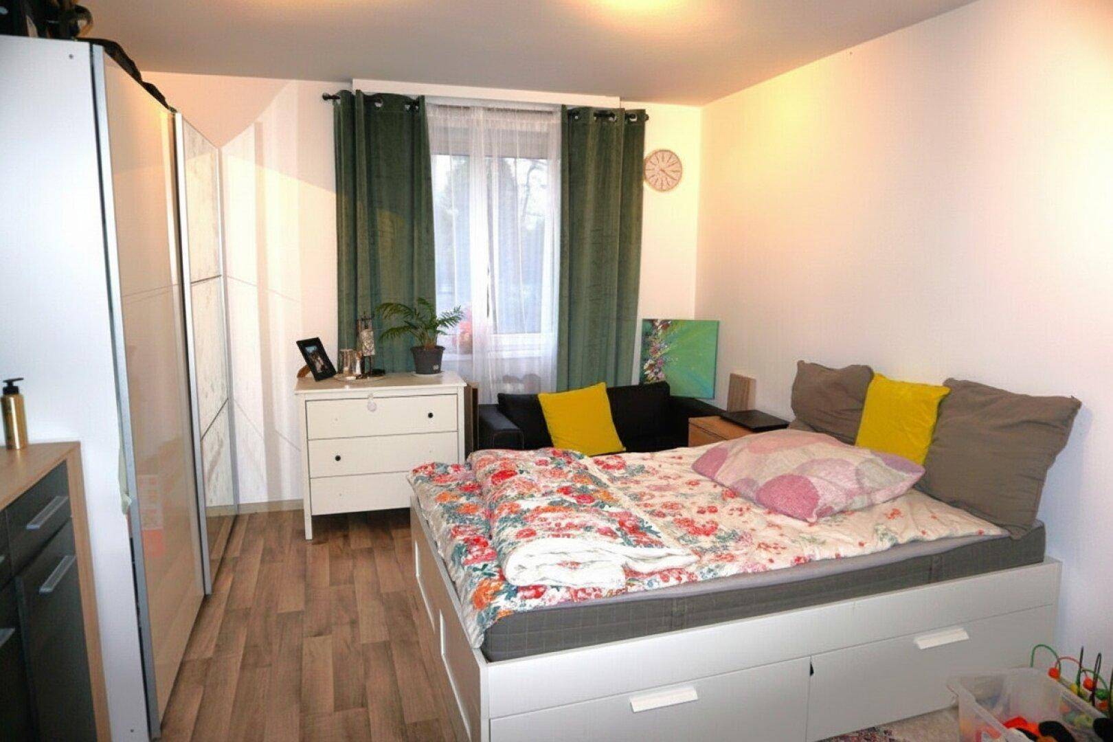Schlafzimmer mit Bett, Kleiderschrank und Fenster, das viel Tageslicht hereinlässt.