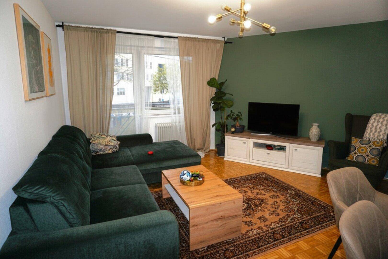 Gemütliches Wohnzimmer mit dunkelgrünem Sofa, Parkettboden und großem Fenster.