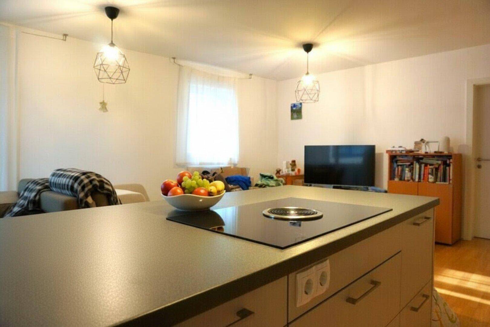 Kücheninsel mit Kochfeld im offenen Wohnbereich, dahinter ein gemütlicher Wohnbereich mit Fernseher.