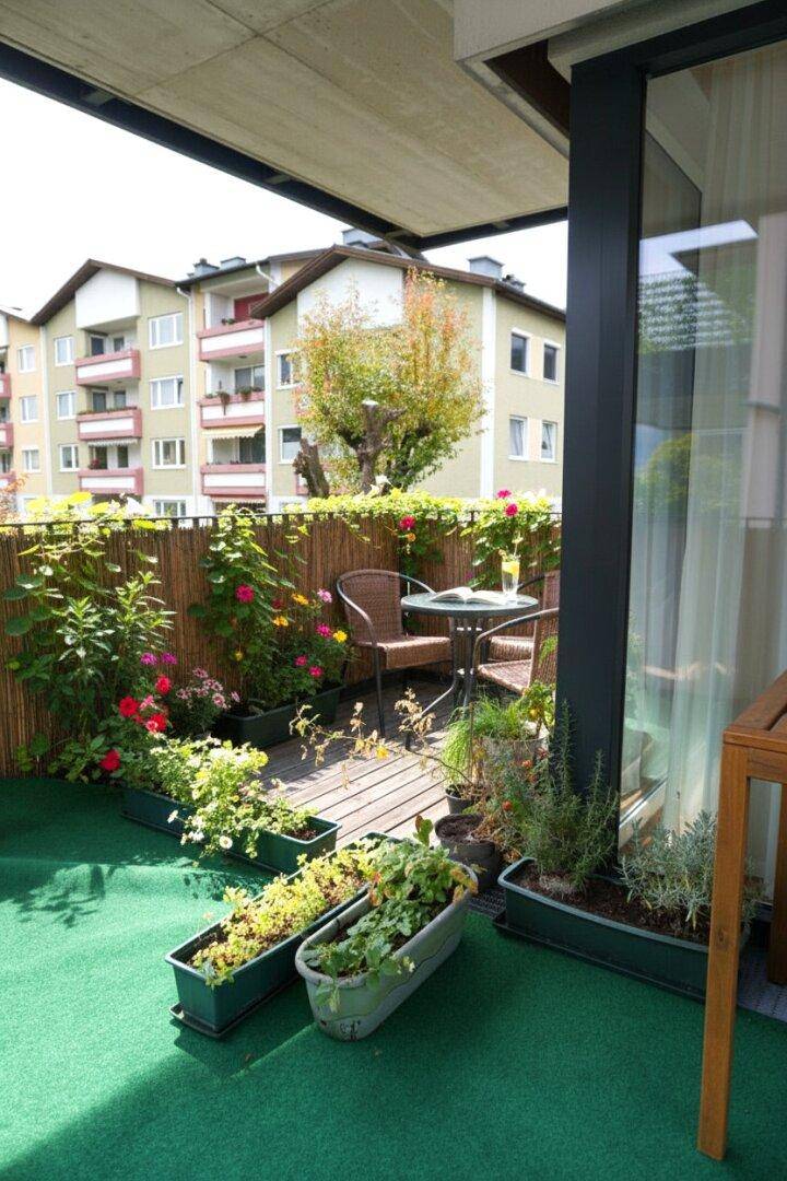 Begrünter Balkon mit Kunstrasen, Holzdeck und kleiner Sitzecke, umgeben von zahlreichen Pflanzen.